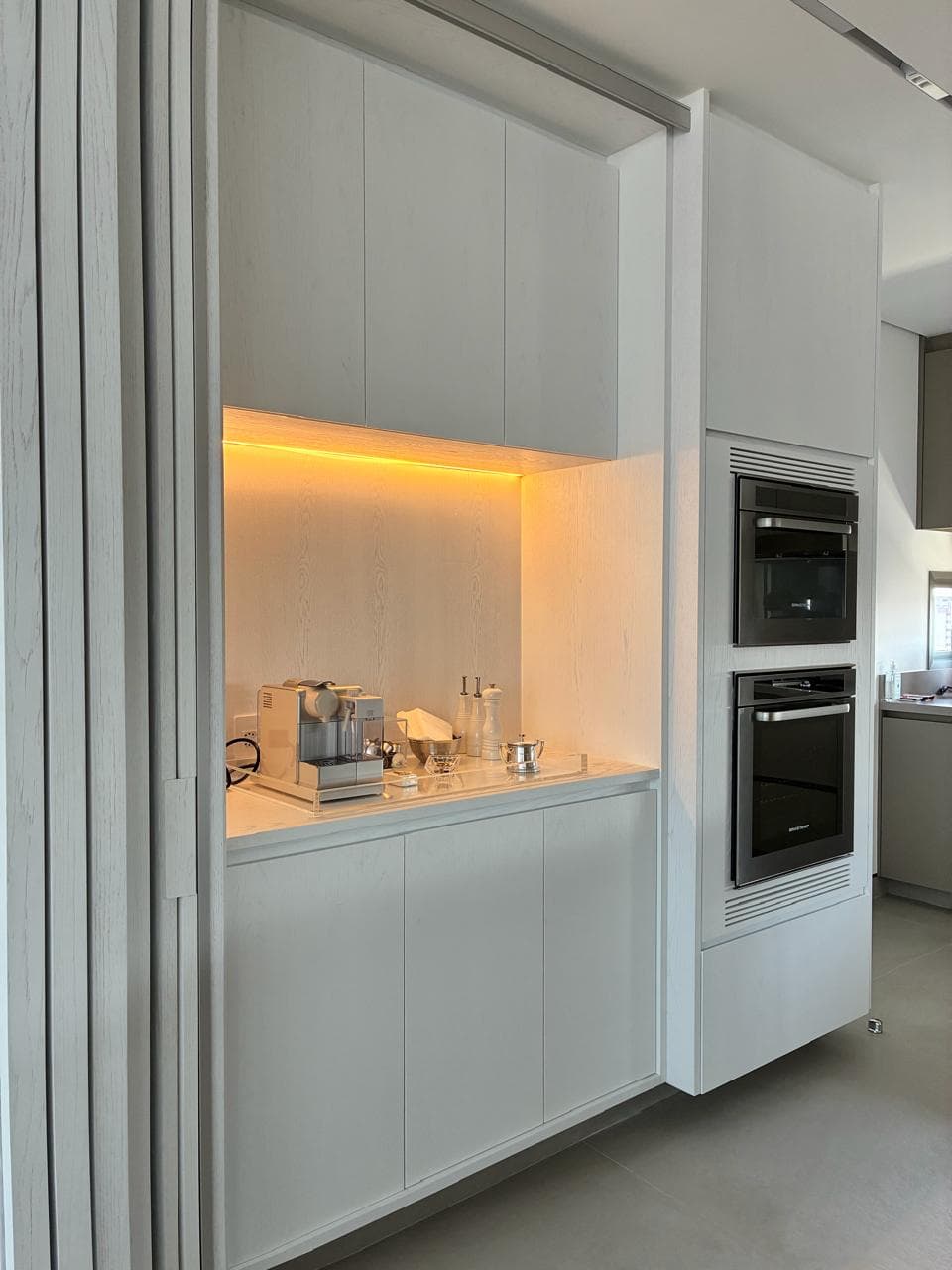 A white kitchen with a coffee station in a warmly backlit niche, handleless fronts and a built-in double oven column.
