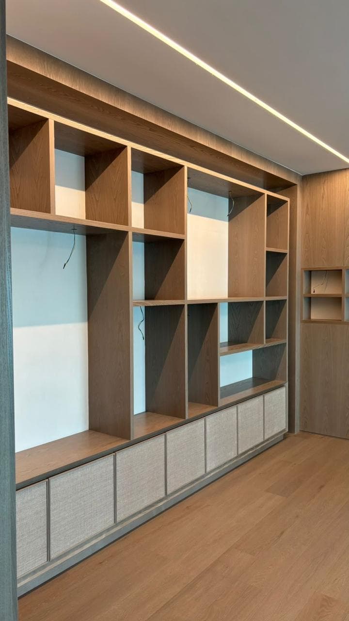 A full-width oak wall unit with open shelving above and fabric-fronted base cabinets below, washed by a ceiling light line.